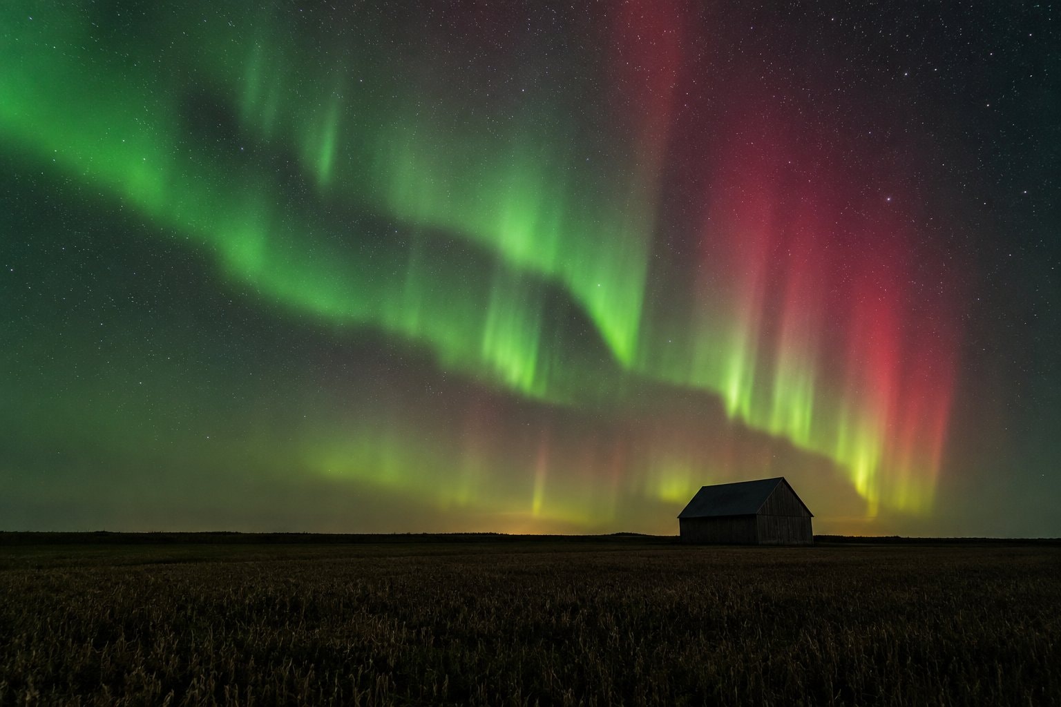 Green and red aurora light above a flat horizon and a distant barn.